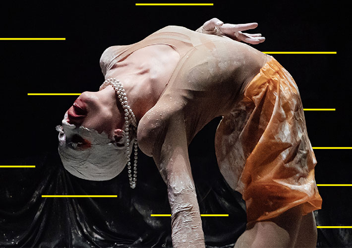 A white woman bending backwards; we see her body from knees up. She wears a stocking bodysuit matching her skin colour and short orange baggy pants, she is stained in plaster. Her face from nose to forehead is all covered in plaster and her lips painted in red cover part of her cheeks. She also wears a white pearl necklace hanging from her neck. There are yellow stripes on the black background.