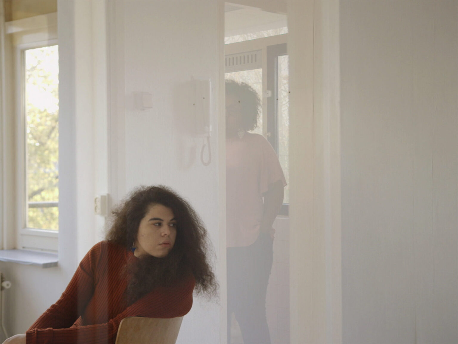 A femme presenting person with long dark curly hair wearing a rust coloured jumper sits in a wooden chair in a bright room. A figure with brown skin and tight dark curls is visible in reflection on a panel behind them.