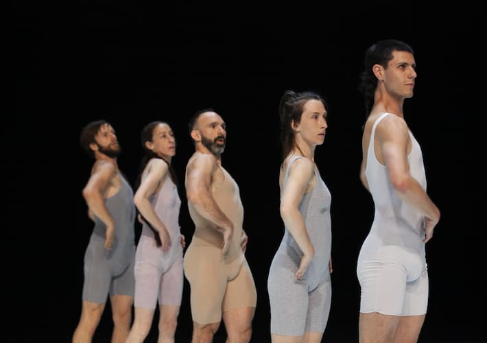 A colour photo of five dancers all wearing neutral color unitards, standing in a vertical line with matching poses. The background is black.