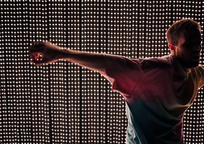 A colour photograph of a white man wearing a tie-dye shirt dancing in front of string white LED lights.He is backlit.
