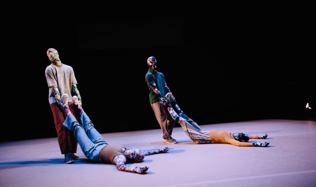 There are four dancers all wearing full-body floral morph suits. Two dancers are positioned on the floor, while the other two dancers are being dragged by the two standing dancers. We see that are on the floor are being positioned into an L shape by the dancers that are standing.