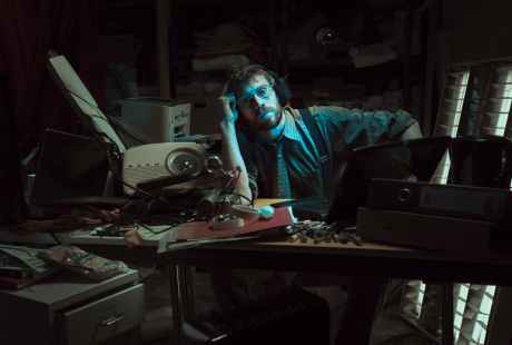 A colour photo of a white man with brown hair who is wearing glasses and is looking deadpan into the camera. He sits at a desk in a room with, vintage computers from the 90s with a light cast on his face.