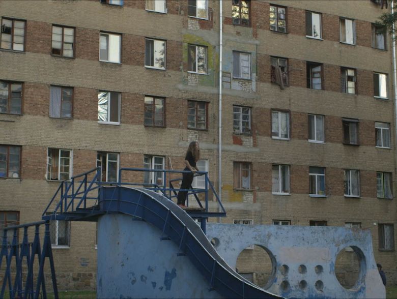 A femme presenting person stands on a blue painted modernist structure in front of what appears to be a housing estate.