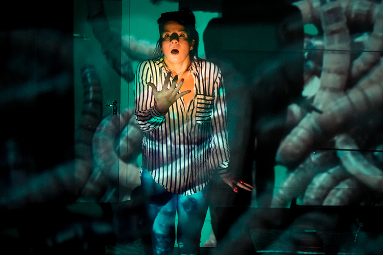 A coloured photo of Marcela Bannitz wearing blue and white striped shirt and blue trousers. She brings her hand almost to her mouth in an expression of astonishment. She is surrounded by worms that look like tentacles designs on canvas.