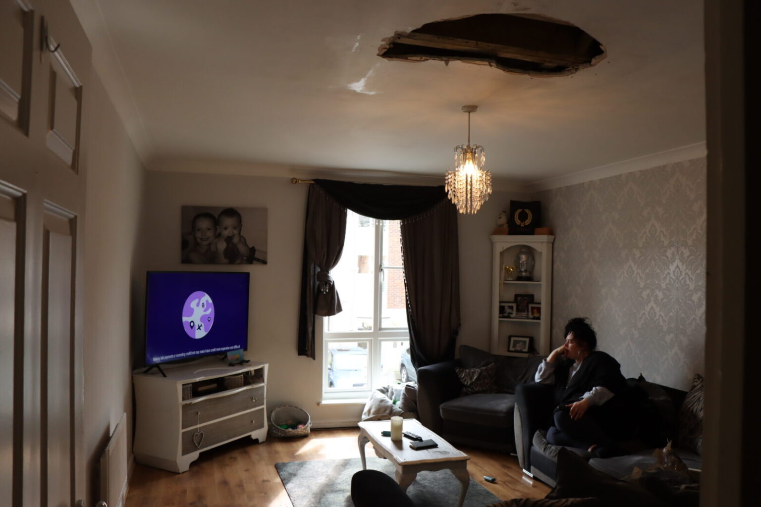 A femme person with dark hair in a pony tail sits watching television in a sitting room with cosy furnishings and sunlight shining in through the window. On the ceiling, beside the light fixture, an enormous dark hole in the plaster exposes the beams behind.