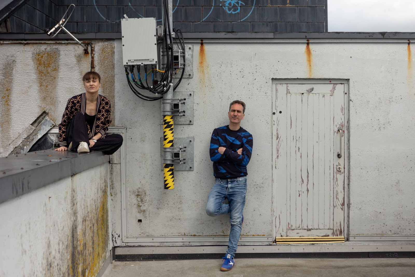 A black man dancing outside in a courtyard facing us. He is looking down and to the left, his legs are crossed, and his arms are out to the sides in a T shape. He is wearing a black and white hat, a light colored button up shirt with a black shirt under it, tan pants, and black shoes.