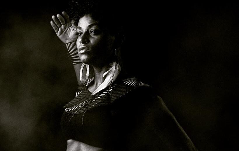 A Black woman in a black and grey photo looking to the right and her torso is on a forward right diagonal. She has her back arm raised and bent at the elbow with the hand up by the head and her front arm is curved low behind her. She is wearing one large circle earring, a woven top and fringe skirt. Her torso is exposed. She appears content.