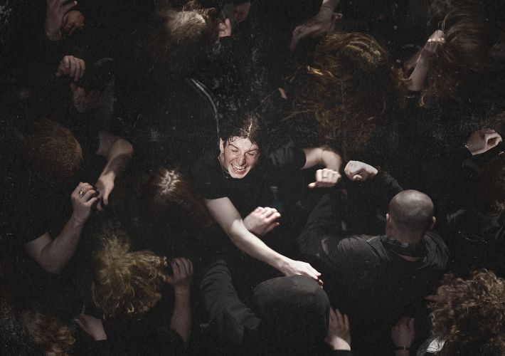 A photo where the viewpoint is looking down from above into a mosh pit of tightly packed people and focusing on one person’s face tilted upwards, eyes closed, mouth open in excitement and arms flailing.
