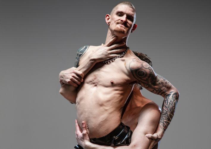 Chris Owen is lifting Dan Daw from behind, who is facing out towards the camera. Both are topless. Dan has tattoos down the whole of his right arm and torso. He has a ginger shaved head and a goatee. Chris has a hand splayed across Dan’s throat, his face hidden behind his back.