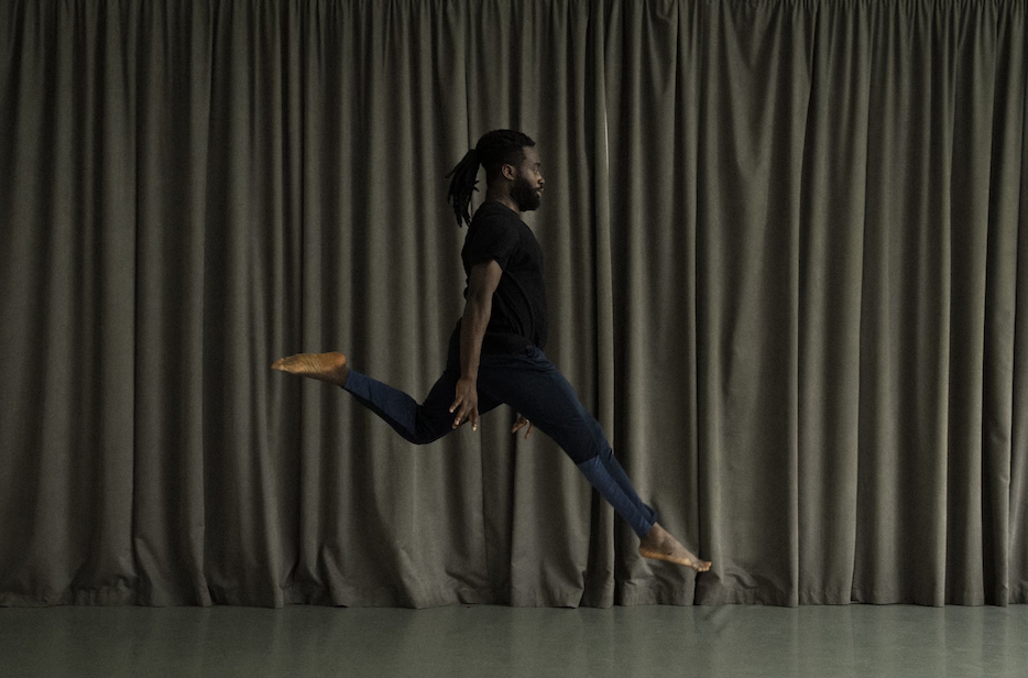 A black man jumping with one leg extended straight to the front and one leg bent to the back. He is wearing a black tee shirt and tight dark blue and black pants. He has a beard and his hair is pulled back. He is facing sideways. Arms are extended straight down by the sides of his body.