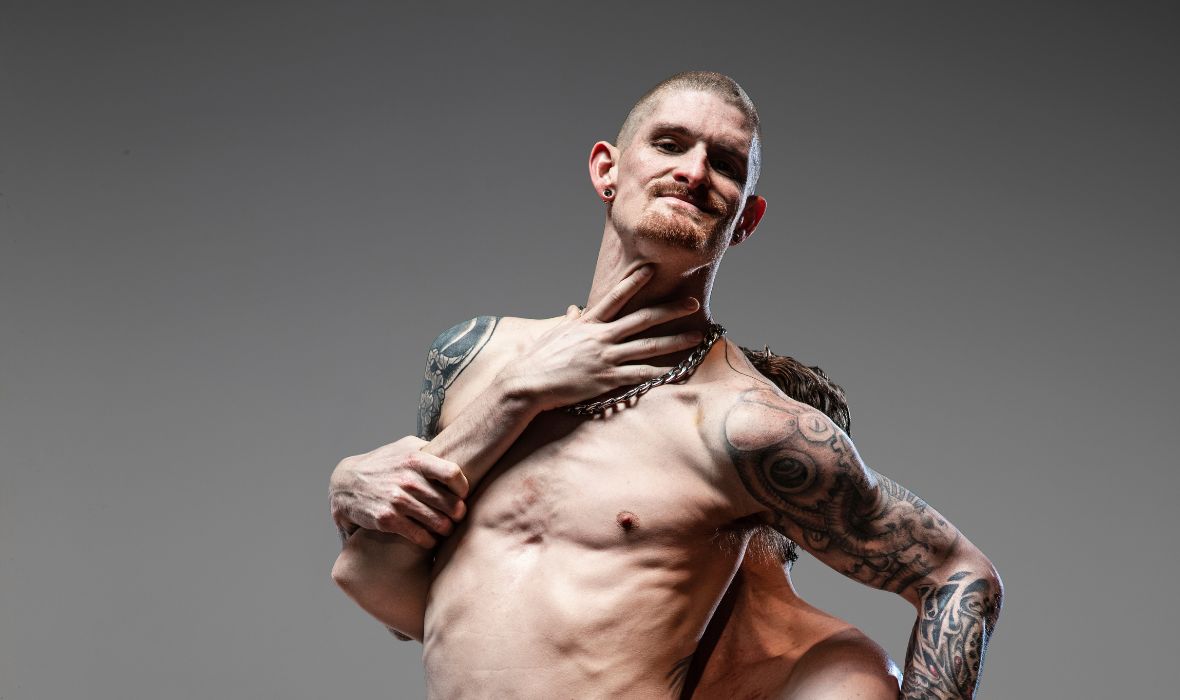 Chris Owen is lifting Dan Daw from behind, who is facing out towards the camera. Both are topless. Dan has tattoos down the whole of his right arm and torso. He has a ginger shaved head and a goatee. Chris has a hand splayed across Dan’s throat, his face hidden behind his back.