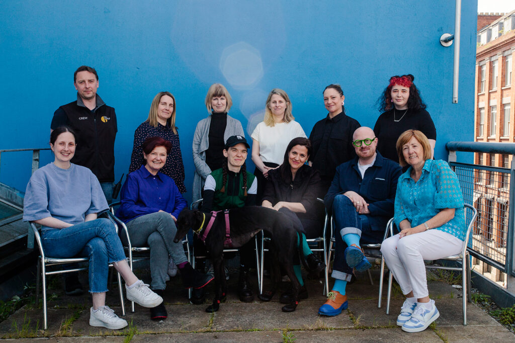 A group of smiling people and a dog sit and stand in front of a blue wall.