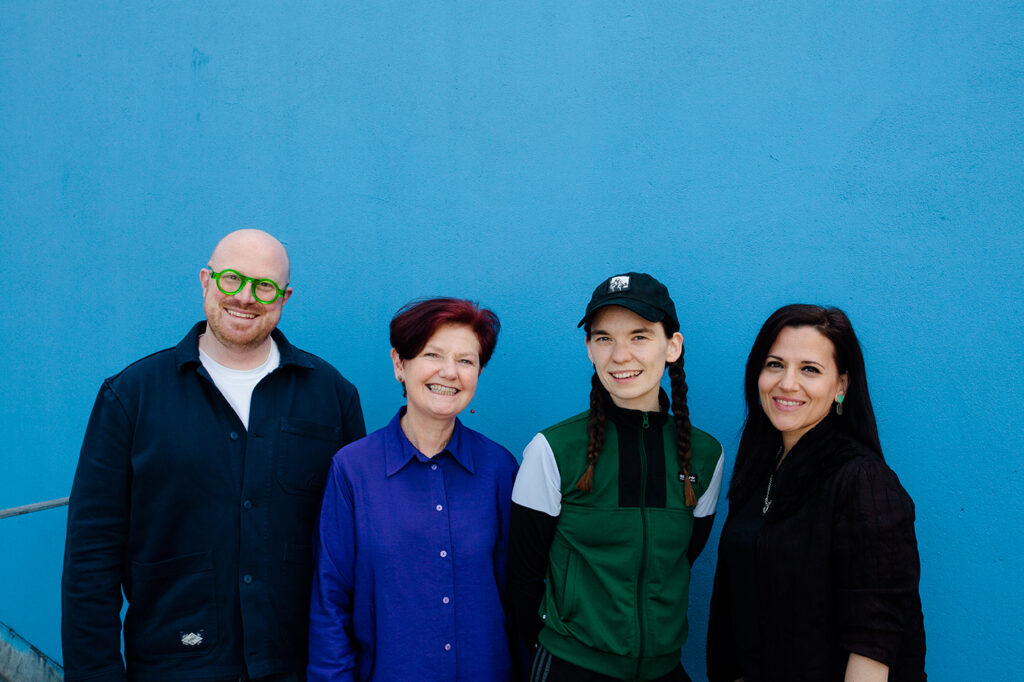 A group of four people stand in front of a blue wall. They are Cian O'Brien, Artistic Director; Orla Moloney, Executive Director; Eimear Walshe, Artist; Sara Greavu, Curator of Visual Arts and they are dressed in various shades of green, blue, black and white.