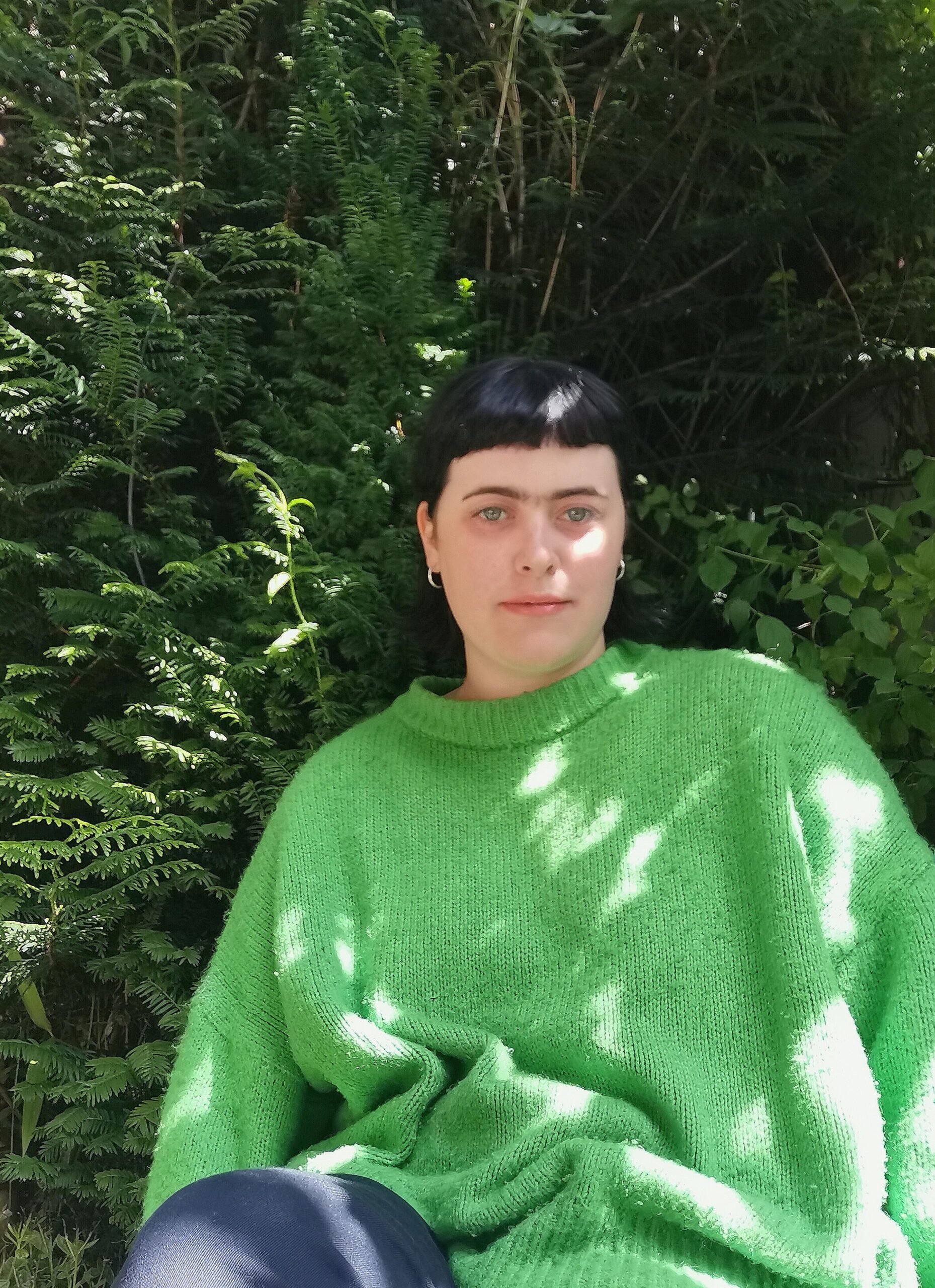 A photograph of Carmen, a white woman in her twenties, poses in her garden. She is wearing a green jumper and is surrounded by foliage. She is looking into the camera.