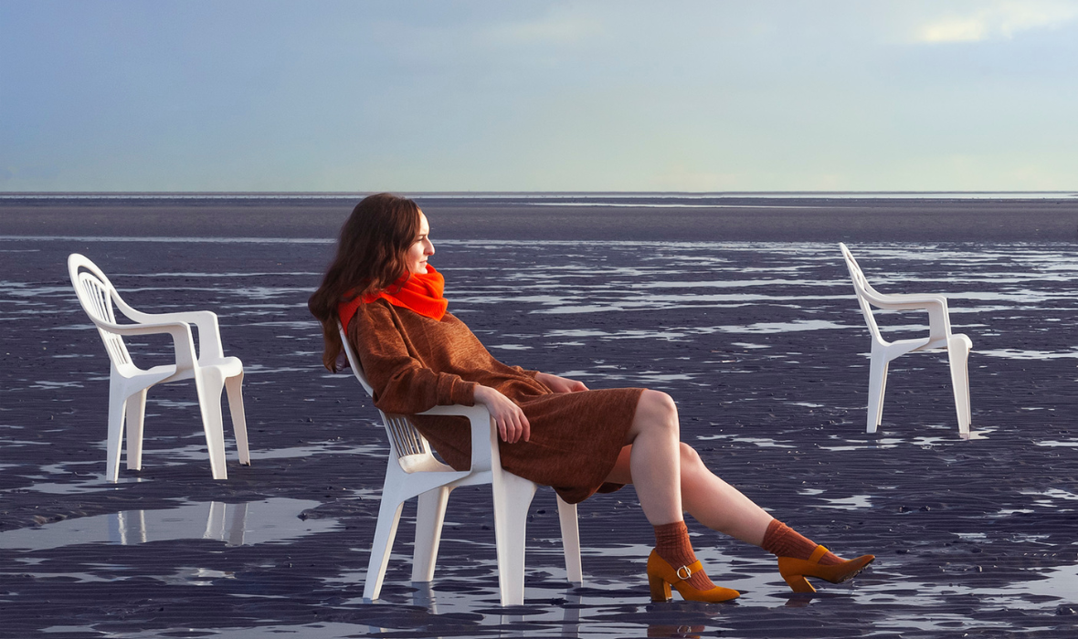 A woman dressed for autumn sits on a white plastic chair on the beach.