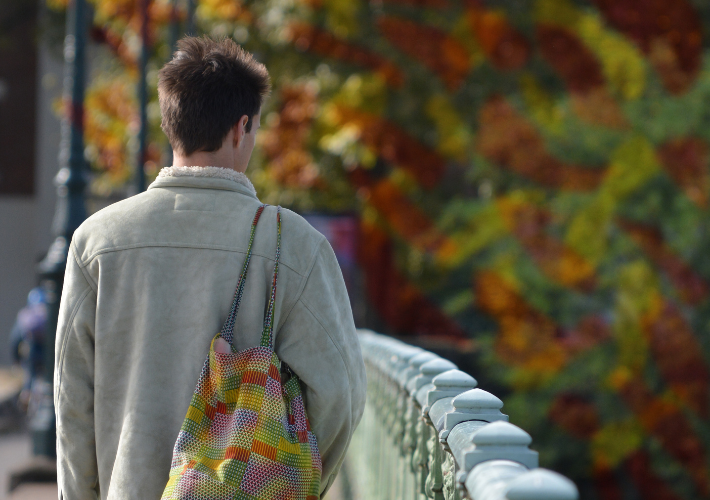 View from behind of a young man wearing a coat walking across a bridge with a bag slung over his shoulder which has the same colour and pattern as the out of focus trees in the background.