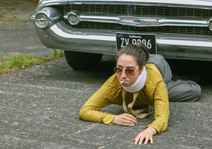 A woman with a cigarette clamped in her mouth and wearing a neck brace and a resolute expression crawls on the road in front of a car.