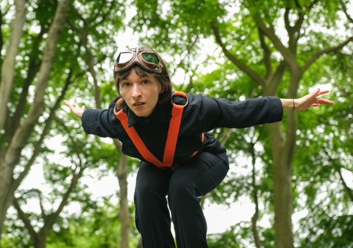 A person wearing a dark pilots outfit and goggles is bending down with arms outstretched as if to fly.