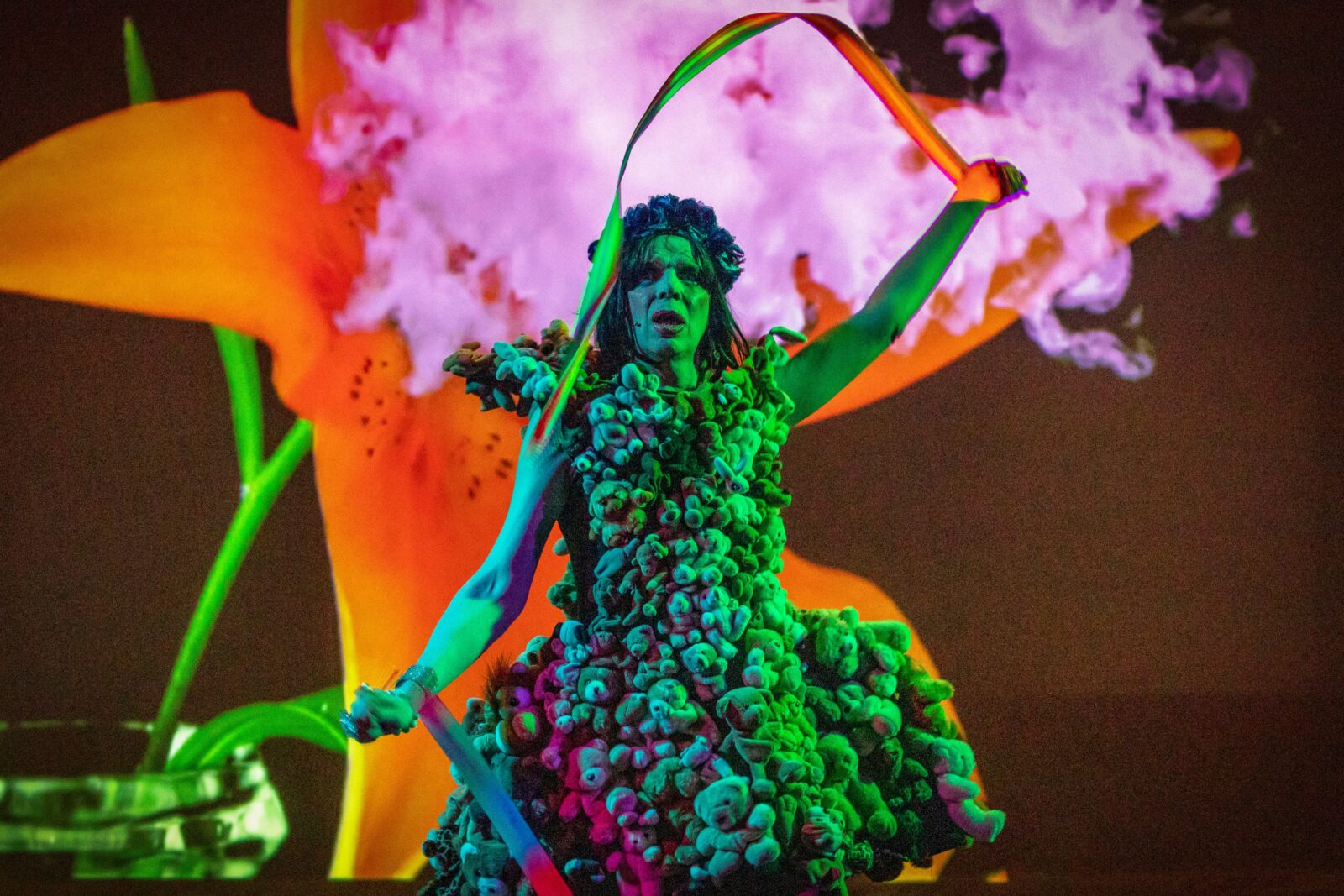 David Hoyle is under a green spotlight. He wears a dress made of Teddy Bears and holds two ribbons. His right hand is above his head. He stands in front of a projection featuring a large orange lily in a glass vase.
