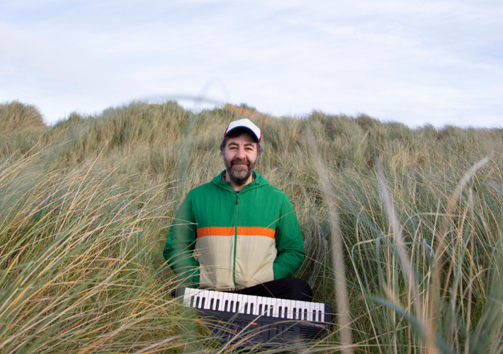 Comedian David O’Doherty standing in tall grasses with a keyboard.