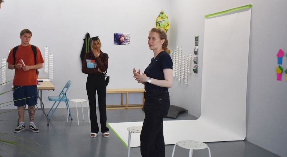 A photo of two Art Walk participants and a guide in a gallery space. There are various stools and chairs throughout the space, a white photoshoot backdrop on the right, and various white accordian-folded pieces of paper hanging on the walls. There are also some coloured pieces of paper on the wall on the right, photographs on the walls, a Hawaiian shirt hanging in the background from the ceiling, a garment hanging on the back wall, and a plant positioned to the left.