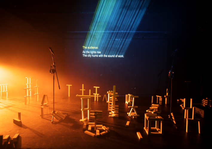 A darkened stage set, with a mic and wooden pieces scattered about, text is projected in light at the back of the set: ‘The Audience: As the lights rise. The city hums with the sound of work’.