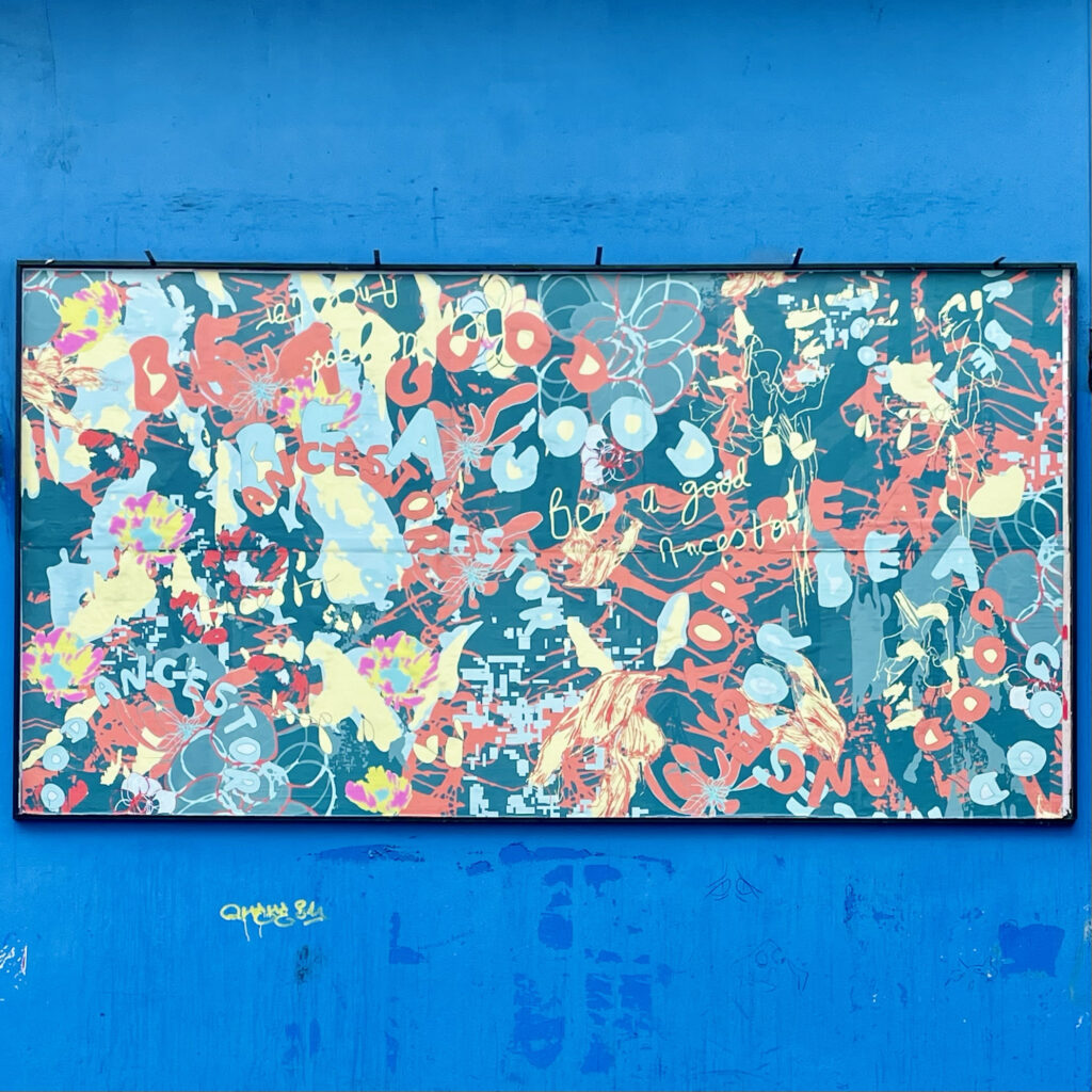 A photo of the billboard on the blue exterior wall of Project Arts Centre. The billboard is a graphic artwork, with the colours yellow, red, and light blue on a teal background. The words ‘Be a Good Ancestor’ appear in circles and swirls, in several placements and sizes across the work. The effect is a vibrant pattern.
