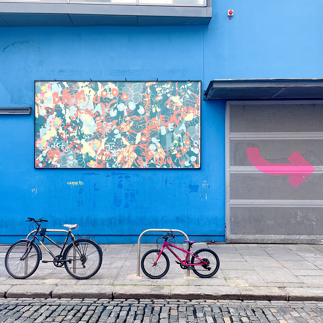 A photo of the billboard on the blue exterior wall of Project Arts Centre. The billboard is a graphic artwork, with the colours yellow, red, and light blue on a teal background. The words ‘Be a Good Ancestor’ appear in circles and swirls, in several placements and sizes across the work. The effect is a vibrant pattern. In front of the billboard is the pavement and the cobbled street, with the arrow to the door of Project in pink on the right. On the pavement, in front of the billboard, an adult bike is locked on the left and a children's bike is locked on the right.