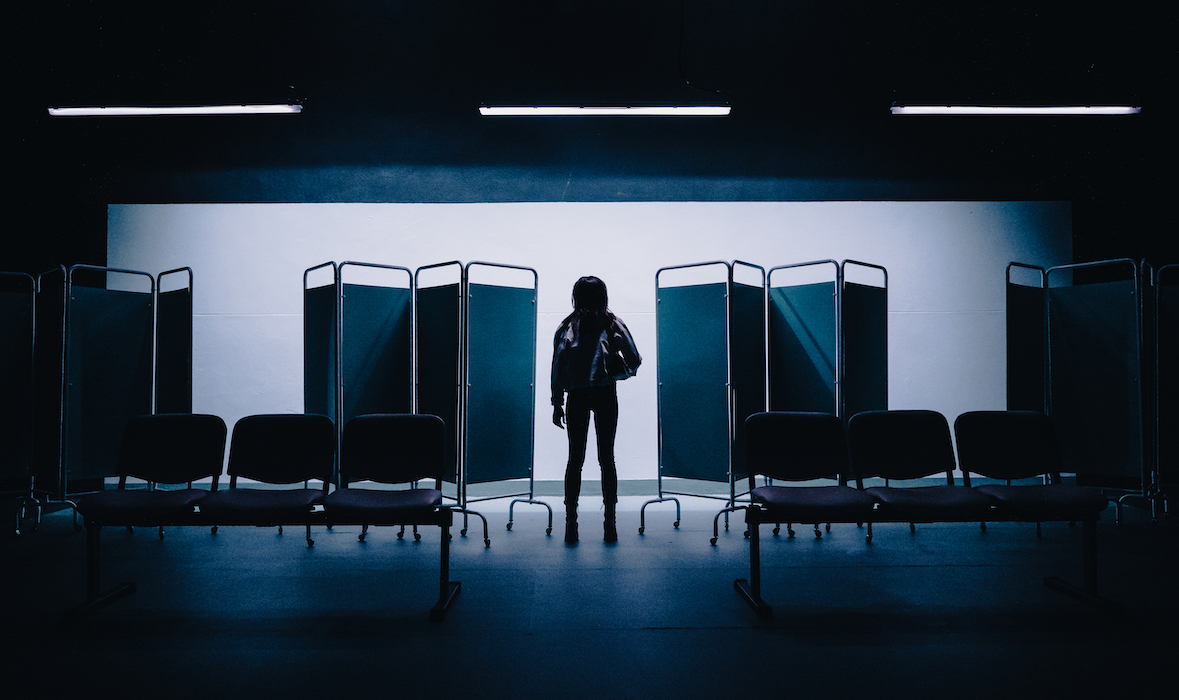 A white back wall. Three fluorescent lights hang from the ceiling. In silhouette, four hospital screens with a woman standing in the centre. The woman has her back turned, face forward, one arm hanging beside and the other holding what looks like an over-the-shoulder bag. Also in silhouette, two sets of hospital seating. Three chairs in each set.