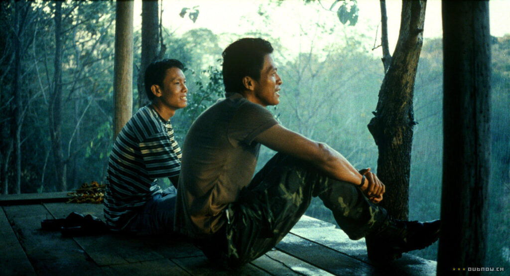Two Thai men sit together on a wooden platform, looking out over a jungle. The one on the left wears a striped t-shirt and jeans. The one on the right wears a military uniform made-up of a khaki t-shirt, camo trousers. and combat boots. Both of them are smiling.