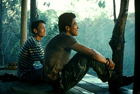 A photo of two Thai men sitting on a wooden platform together looking out over a wooded jungle. They are both smiling.