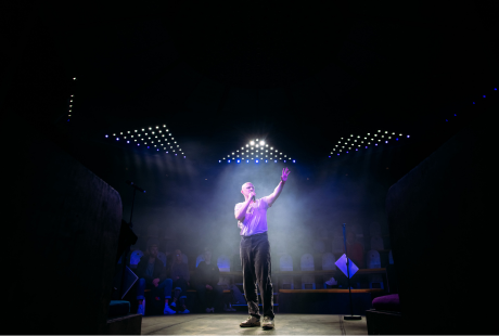 A performer stands stage with one hand holding a microphone, and the other hand raised in the air. An audience is seated behind them, in a circular seating arrangement. The space is darkly lit, and the performer is spotlit in the centre of the stage.