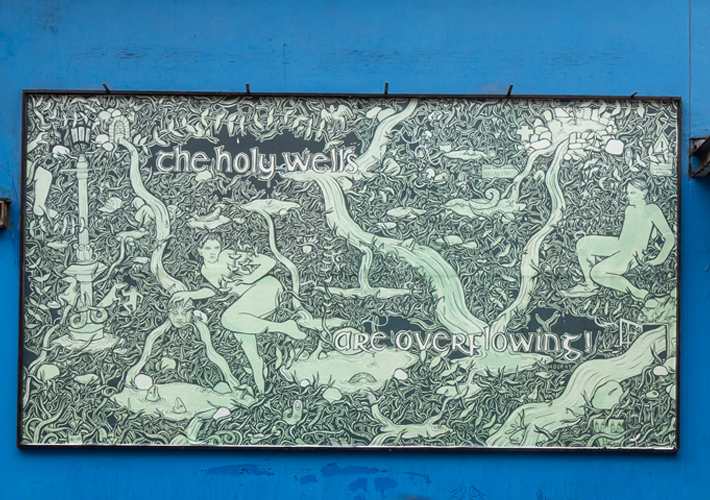 A photo of the billboard on the blue exterior wall of Project Arts Centre. The billboard artwork is a green and black drawing of water overflowing from Holy Wells. Two surprised figures are surrounded by dense foliage. Fish and strange creatures are appearing from the water. Small details in the drawing reveal the setting is Dublin city. The text reads “The Holy Wells are Overflowing! (Hooray)”