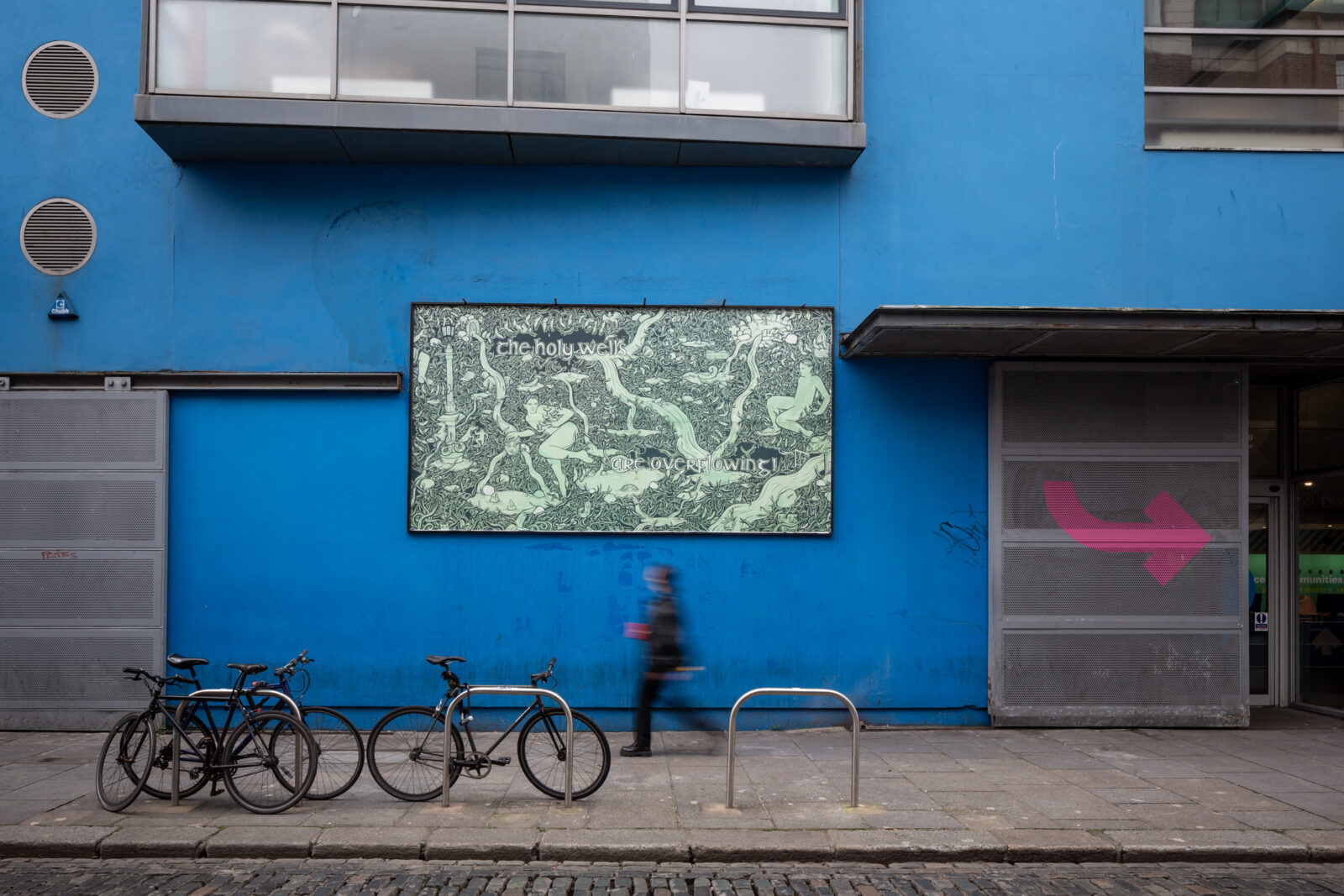 A photo of the billboard on the blue exterior wall of Project Arts Centre. The billboard artwork is a green and black drawing of water overflowing from Holy Wells. Two surprised figures are surrounded by dense foliage. Fish and strange creatures are appearing from the water. Small details in the drawing reveal the setting is Dublin city. The text reads “The Holy Wells are Overflowing! (Hooray)." There is a blurred figure walking on the pavement in front of it and some bikes locked to railings.