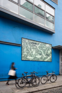 A photo of the billboard on the blue exterior wall of Project Arts Centre. The billboard artwork is a green and black drawing of water overflowing from Holy Wells. Two surprised figures are surrounded by dense foliage. Fish and strange creatures are appearing from the water. Small details in the drawing reveal the setting is Dublin city. The text reads “The Holy Wells are Overflowing! (Hooray)." There is a blurred figure walking on the pavement in front of it and some bikes locked to railings.