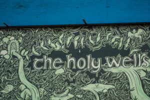 A close-up of an artwork detail on the Project billboard. Text reads "The Holy Wells" against a green and black drawing of water overflowing, dense foliage and fish. On the left is a cap which reads "Kiss Me I'm Irish."