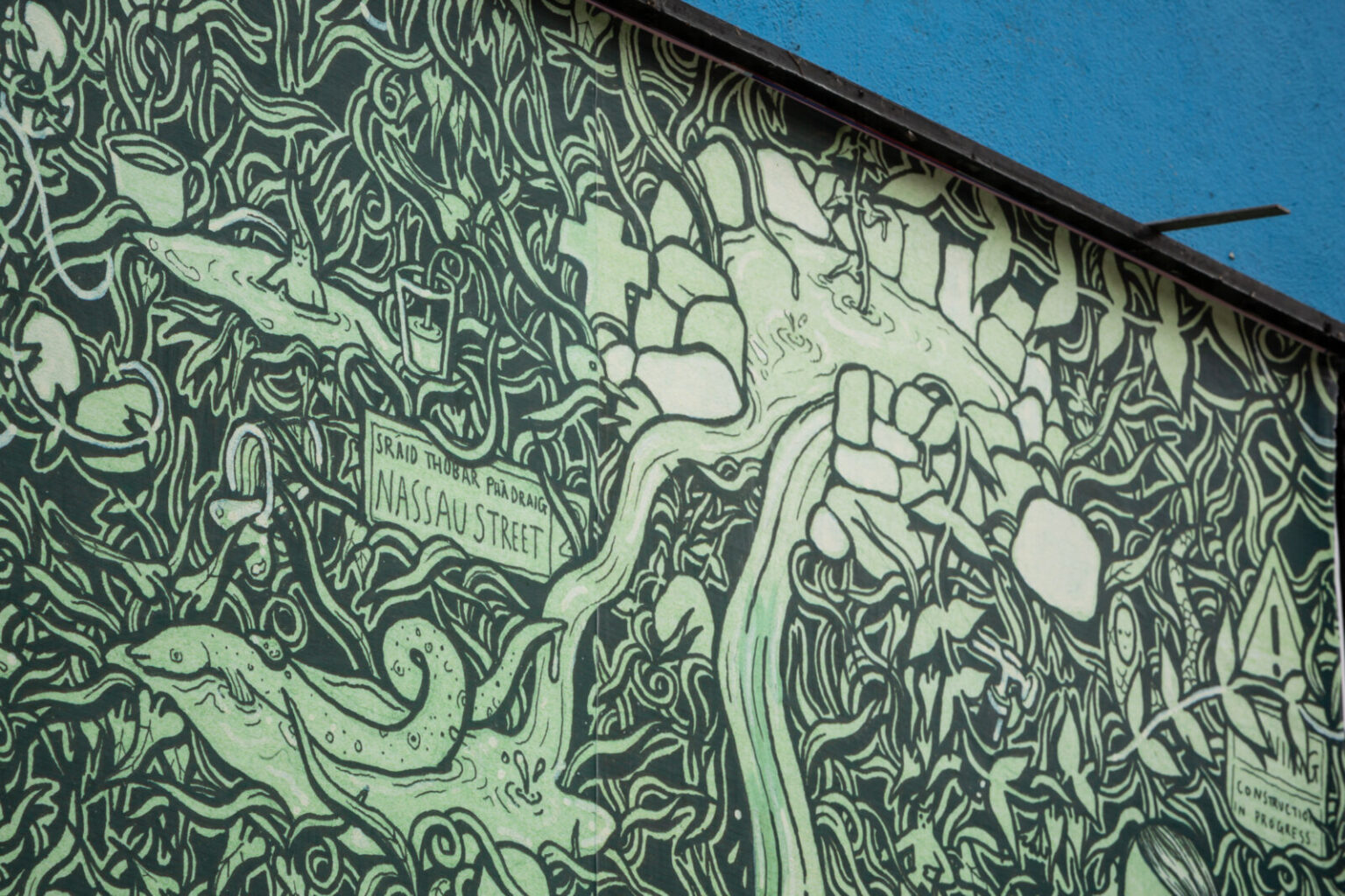A close-up of an artwork detail on the Project billboard. The artwork shows a green and black drawing of water overflowing from Holy Wells. Fish and strange creatures are appearing from the water. Nestled amongst the foliage are two signs, a street sign for Sráid Thobar Phádraig/Nassau Street, and a warning sign which reads “Warning. Construction in Progress”