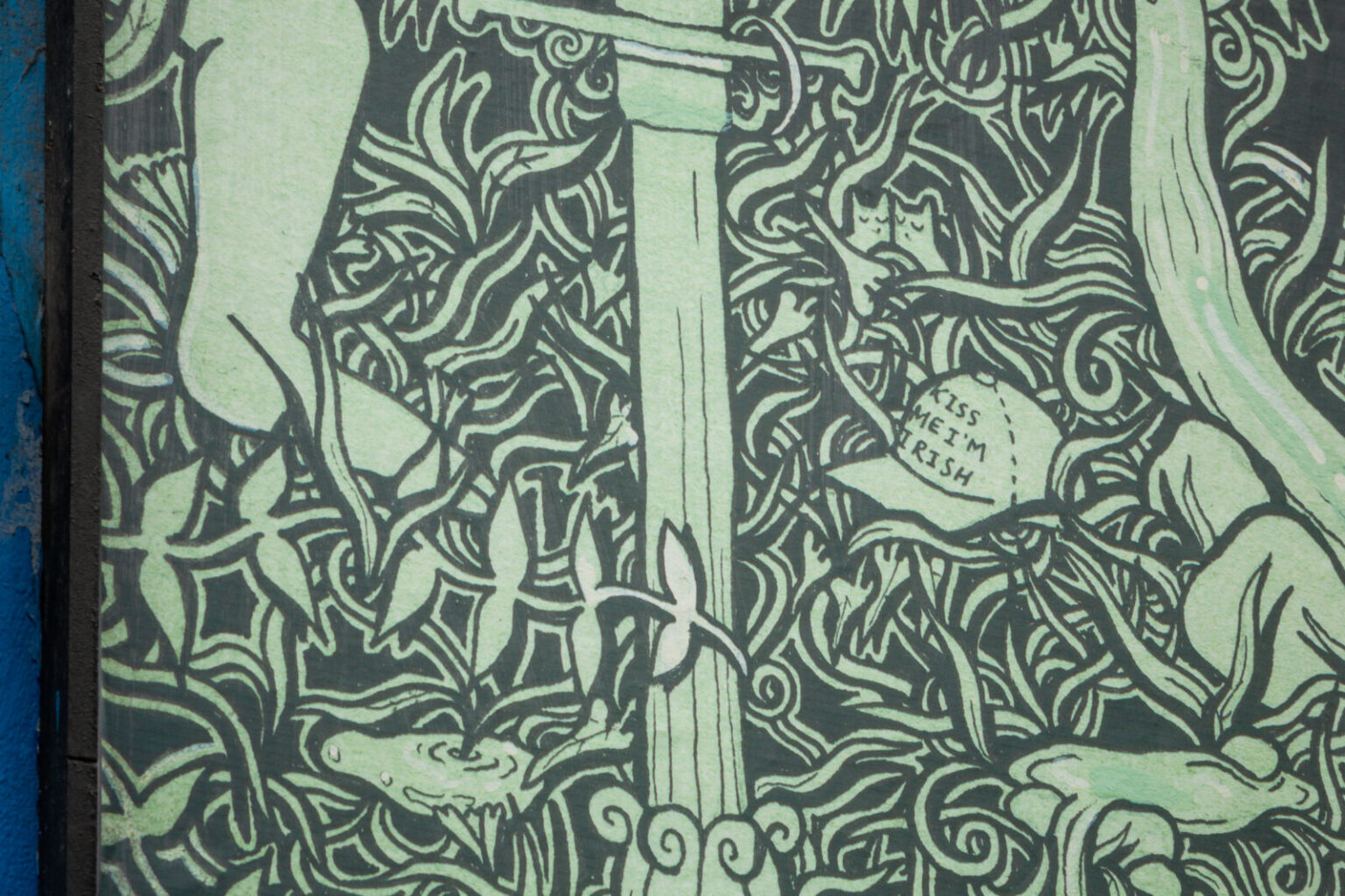 A close-up of an artwork detail on the Project billboard. The artwork shows a green and black drawing of water overflowing from Holy Wells. There is a section of a lamppost pillar nestled in dense foliage in the centre, and a cap sitting amongst the leaves on the right which reads “Kiss Me I’m Irish.”
