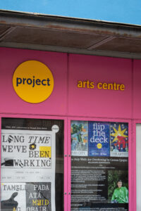 A photo of the first two pink poster boxes at the front of the Project building. The posters are for exhibition Long Time We've Been Working and for Carmen Quigley's Whip It Up! Flip the Deck billboard commission the Holy Wells Are Overflowing!