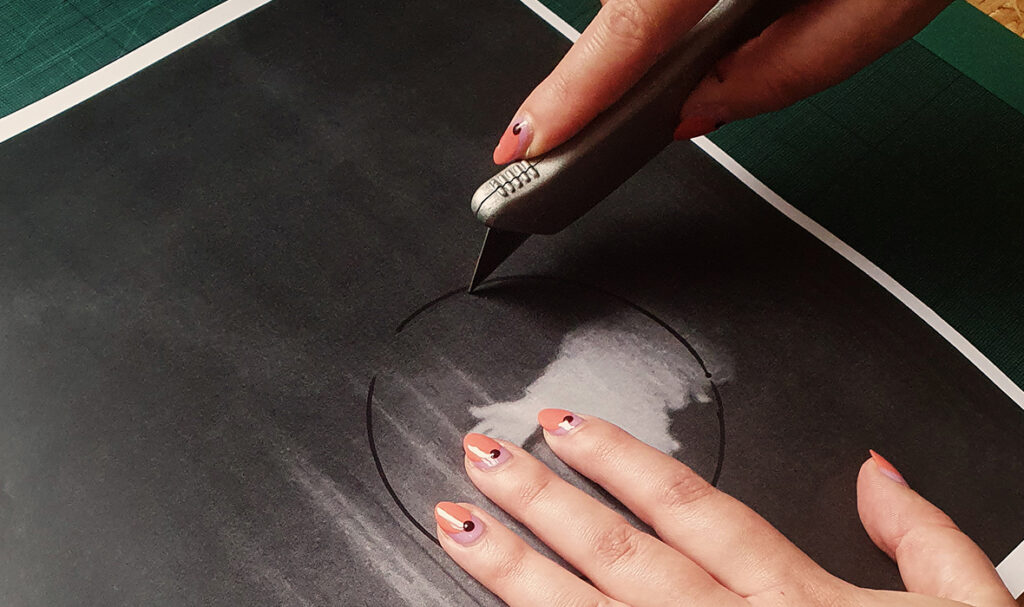 Image of hands at work, cutting a section of an image from a black and white photocopy. The hands have pale skin and nails painted with a design in peach and black. They hold a craft knife and brace the photocopy on a green cutting mat on a table of OSB. A Sharpie marker is visible to the side.