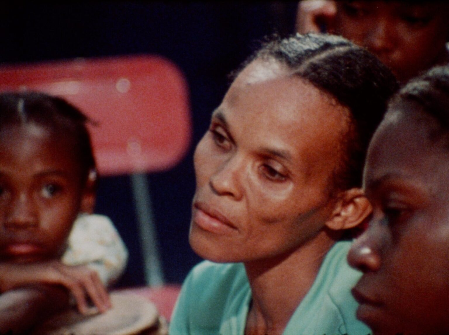Centred in the photo is a closeup of a woman wearing an aqua green sweatshirt. She has medium brown skin and wears her hair parted in the middle and pulled back. Her cheekbones are impossibly high. In the foreground, just seen, is a woman in profile with darker skin. In the background, a young girl looks on with her chin resting on her hands. A red chaire can be seen behind her.