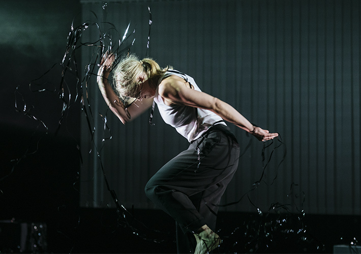 Sibéal Davitt, a white Irish woman in her thirties, is dancing alone on a stage. The image showcases the dancer in profile, mid-dance in a dynamic pose against a dark background. Clothed in a lighter-toned sleeveless top and darker trousers, the dancer's body is slightly twisted with head bent and arms outstretched, embodying a sense of movement, excitement and power. One knee is raised in a jump, as if she is about to stamp her foot. The darkness of the background accentuates the energy in the scene. Strands of black foil are caught in her hands and tossed in the air around her, catching the light.