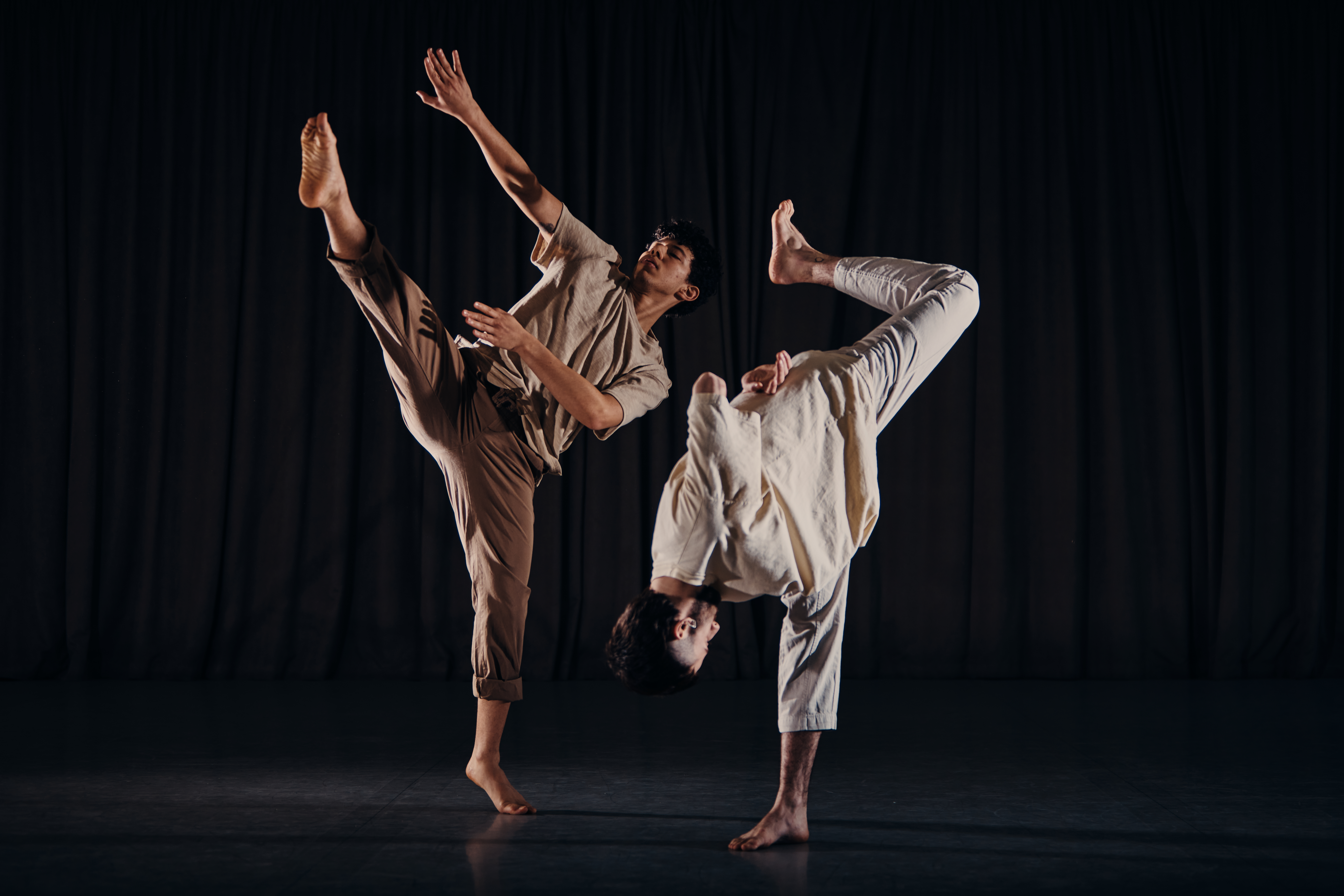 On the left, a male dancer in partial relevé on his left leg, his right leg is raised as far as it can go towards the house left. His torso leans towards the male dancer on the right. His shoulders and face are relaxed, but his right arm is raised and parallel to his right leg. His left arm relaxes towards the ground, but bends so that his forearm is parallel to both his right arm and leg, landing between the two. On the right, another male dancer stands on his right foot, folding himself in half. His arms are tucked behind his back and his left leg is raised hight in the air and bent at a 90° angle.