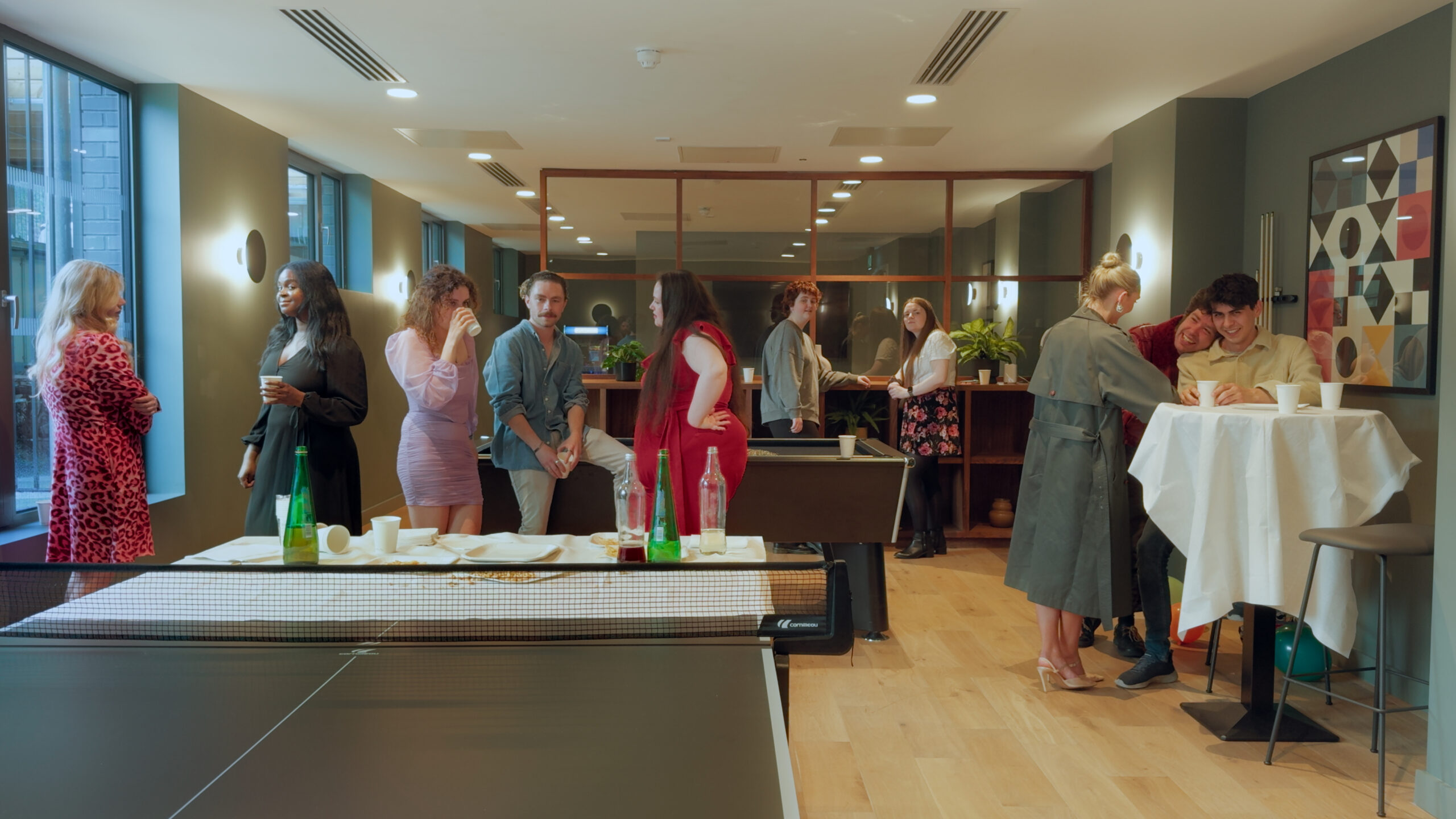 The image shows a scene of ten young people in a space decorated in a corporate, ’tasteful’ style. The colour palette is muted green and warm wood tones. They are in pairs or small groups talking and there is food and drink visible around them, including cups, plates and bottles on a pool table and ping pong table in the foreground.