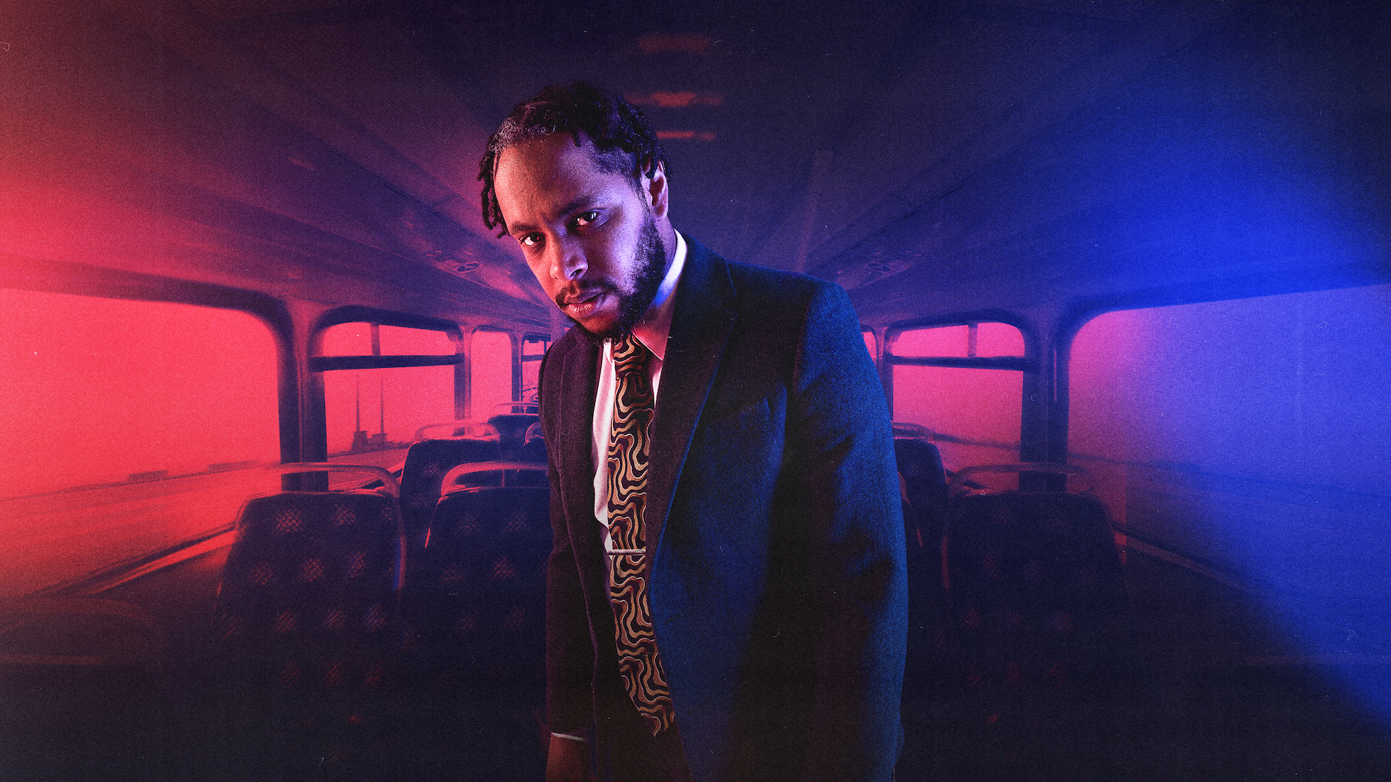 A solitary figure, the writer and performer Kwaku Fortune, is pictured upstairs on a Dublin bus, illuminated by red and blue lights on either side. He is wearing a dark suit, and a tie with a yellow and black swirling pattern. He is looking directly at the camera. There is a look of both sadness and anger on his face. The surrounding darkness of the image contrasts with the light, creating a moody and introspective atmosphere. The image evokes themes of solitude, identity, and contemplation.