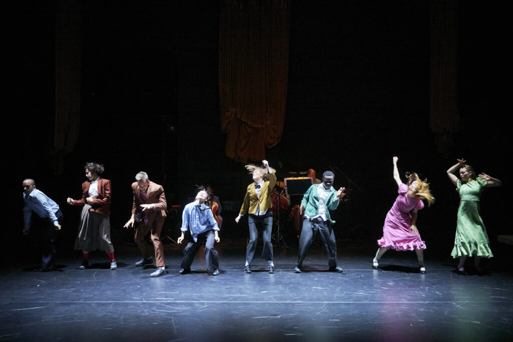 A group of eight performers are captured mid-movement on a dimly lit stage. They are dressed in a mix of colourful costumes — some wearing suits in shades of brown, blue, and green, while others wear flowing dresses in pink and green. Each performer strikes a distinct dynamic pose, suggesting an energetic and expressive dance sequence. The stage backdrop is dark, with two large draped curtains hanging in the background, and musicians and instruments faintly visible behind the dancers. The lighting focuses on the performers, highlighting their movement and expressions.