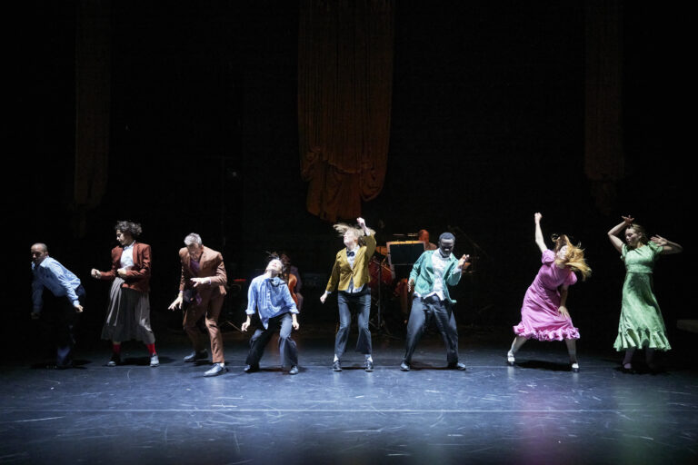 A group of eight performers are captured mid-movement on a dimly lit stage. They are dressed in a mix of colourful costumes — some wearing suits in shades of brown, blue, and green, while others wear flowing dresses in pink and green. Each performer strikes a distinct dynamic pose, suggesting an energetic and expressive dance sequence. The stage backdrop is dark, with two large draped curtains hanging in the background, and musicians and instruments faintly visible behind the dancers. The lighting focuses on the performers, highlighting their movement and expressions.
