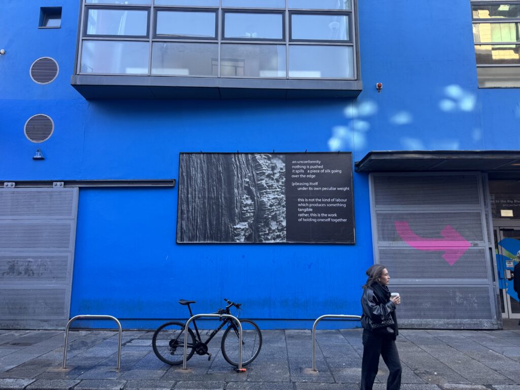 Image description: A billboard situated on a blue wall. On the billboard is a black and white photographic close up of rock strata running vertically meeting a separate rock formation of horizontal rock layers. This is bordered by a paragraph of white text on a solid black background to the right. The text is staggered, appearing all in lower case and contains no punctuation, formally referencing the stratified rock. The text reads: an unconformity, nothing is pushed. It spills, a piece of silk going over the edge (pl)easing itself under its own peculiar weight. This is not the kind of labour which produces something tangible. Rather, this is the work of holding oneself together.