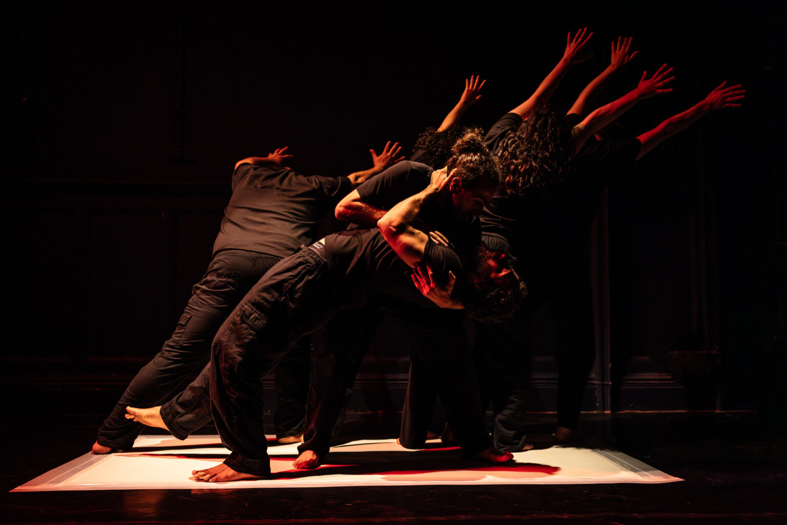 A group of performers dressed in black moves together in a powerful contemporary dance formation on a dimly lit stage. Their bodies lean and reach in unison, creating a sculptural shape under warm red and amber lighting, conveying strength, resistance, and collective movement.