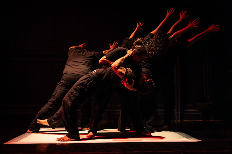 A group of performers dressed in black moves together in a powerful contemporary dance formation on a dimly lit stage. Their bodies lean and reach in unison, creating a sculptural shape under warm red and amber lighting, conveying strength, resistance, and collective movement.